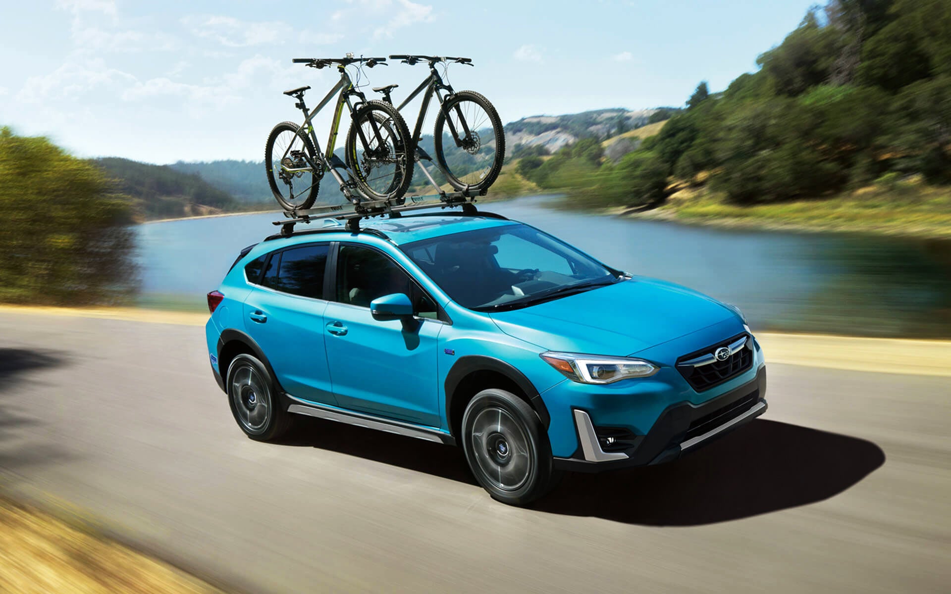 A blue Crosstrek Hybrid with two bicycles on its roof rack driving beside a river | Sommer's Subaru in Mequon WI
